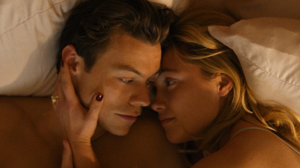 The man, Harry Styles, and the woman, Florence Pugh, lying in bed together, sharing a peaceful moment of intimacy and closeness as they face one another with her hand holding his cheek. 