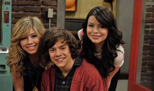 Jennette McCurdy on the left, Harry Styles in the middle, and Miranda Cosgrove on the right pose together smiling on the set of the TV series iCarly. 