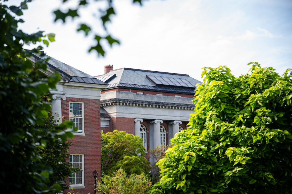 A partial view of classic red brick university buildings, Old Main, with large white columns and arched windows, framed by vibrant green trees under a bright sky.