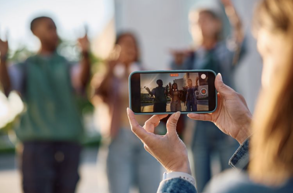 woman - standing almost off screen - holds a phone, taking a video of three people with their hands half raised.