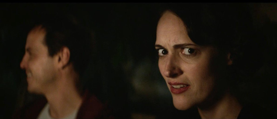 Fleabag and the Priest are in a car. Fleabag is in the camera’s focus and breaks the fourth wall. The Priest is looking in a different direction. Fleabag has a confused and concerned expression.