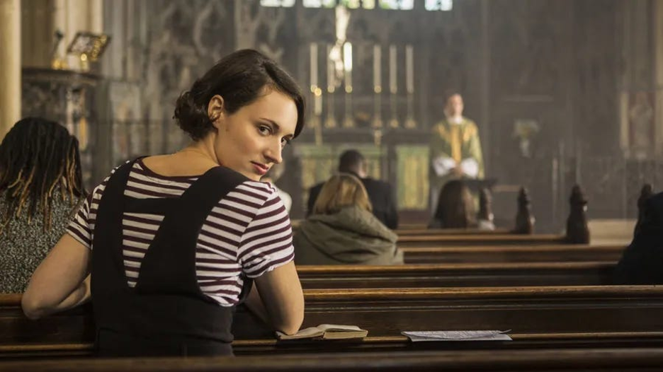 Fleabag is in a church sitting in one of the pews, turning backwards to look at the camera sneakily.