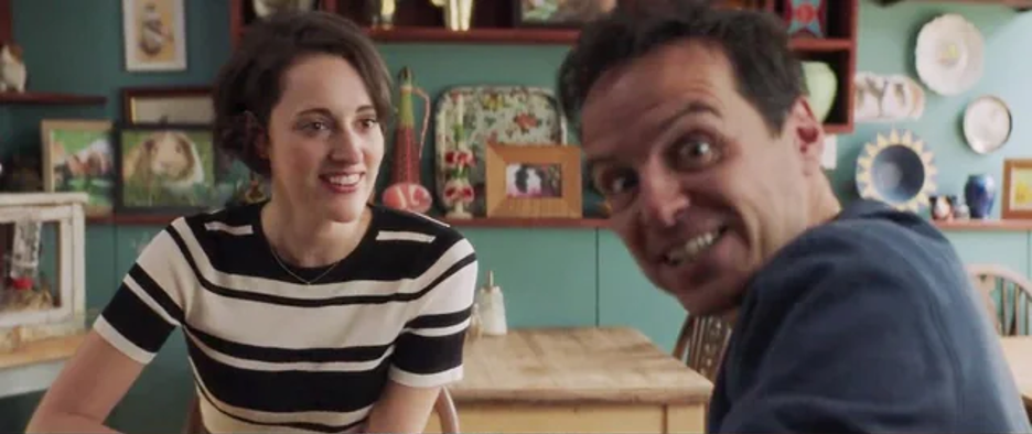 The Priest and Fleabag are sitting at a table in her cafe. The Priest is breaking the fourth wall looking at the camera with a shocked and crazed expression. Fleabag is looking at the Priest seemingly unaware.