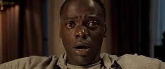 Close-up of a distressed Black man with wide eyes and a tear streaming down his cheek, sitting in a dimly lit room, appearing hypnotized and frozen in fear.