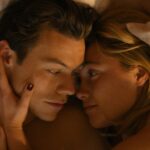 The man, Harry Styles, and the woman, Florence Pugh, lying in bed together, sharing a peaceful moment of intimacy and closeness as they face one another with her hand holding his cheek. 