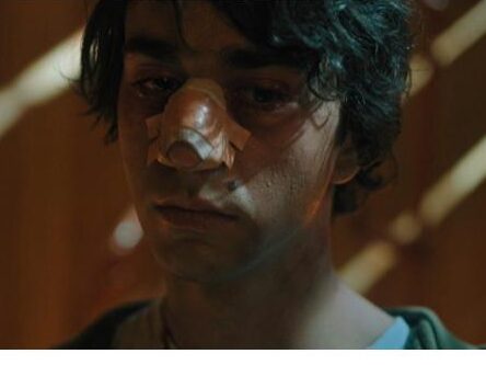 Peter stands with a blank expression and a broken nose covered in bandages after being possessed by the demon Paimon. Warm light surrounds him as he is silently hailed by unseen followers, symbolizing his complete loss of self.