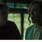 Annie, possessed by the spirit of her daughter Charlie, screams in fear during a failed ritual to make contact with her. Her husband Steve stands nearby, watching in alarm as the possession unfolds.