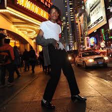 Jordan Neely, dressed as Michael Jackson, poses on a busy New York City sidewalk at night. He wears a white T-shirt, black pants, and sparkling gloves and socks, with bright theater lights, pedestrians, and taxis in the background.
